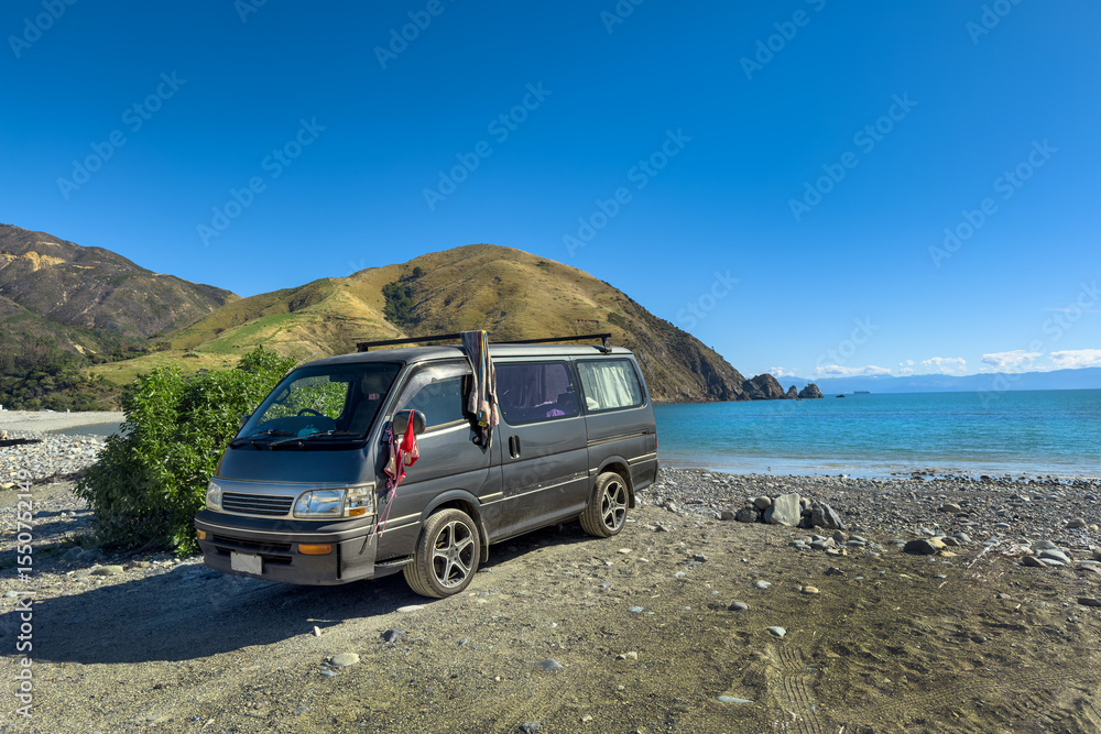 Naklejka premium New Zealand. Tourists' campervan at Cable Bay, Nelson Region. Tranquil South Island beach. Bikini and towel drying on side of van. Sentinel Hill in distance.