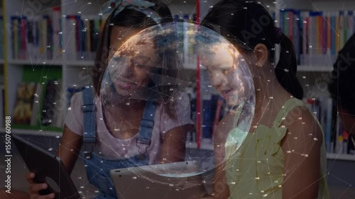 Two preteen girls tapping tablets in library, generating rotating 3D globe for digital learning