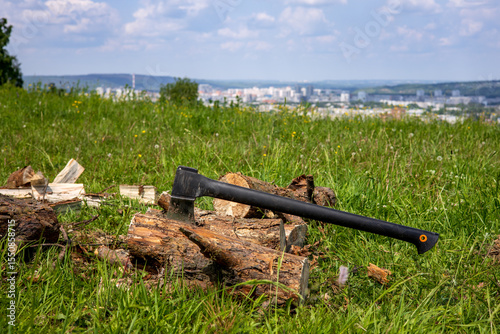 A tourist axe chops firewood for a picnic. Landscape with green grass, blue sky with clouds and a city in the distance.