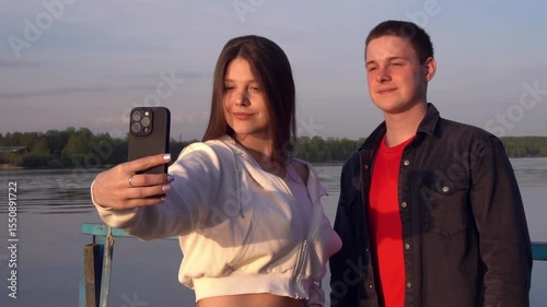 A girl takes pictures of herself and her brother on smartphone, creating summer photos from a trip along the river. young tourists at the river pier.