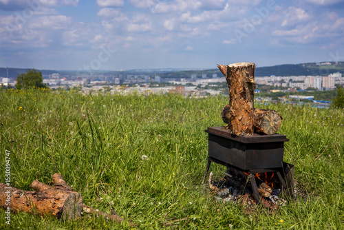 Smokehouse with open fire on grass against the background of a landscape with a city in the distance.