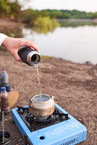 Person pouring water in a kettle to heat it with a travel stove on the bank of a stream, at sunrise. El Palmar National Park, Entre Rios, Argentina.