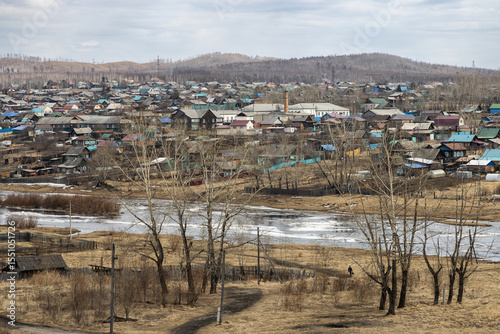 Mogocha city, Transbaikal region, Eastern Siberia, Russia. View of low-rise buildings in a Siberian city. A man is walking along a country road. In the distance are wooden houses and hills. Spring