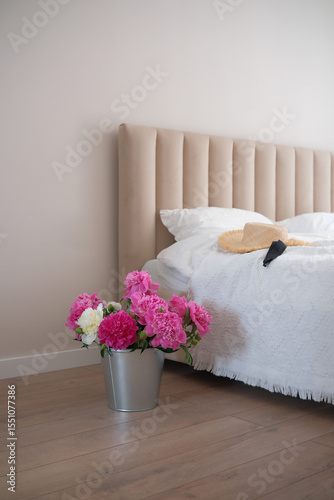 In a cozy bedroom, pink peonies bloom, a comfy bed is nearby, and a stylish hat rests on the bedside table
