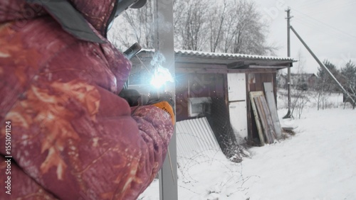 A person dressed in warm winter clothing diligently works on a chilly, snowy day near a rustic wooden structure