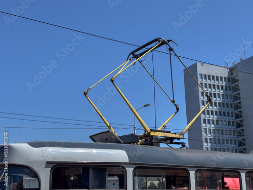 Tram pantograph and overhead lines against a clear blue sky with a modern building, concept of public transport, urban infrastructure, city development.