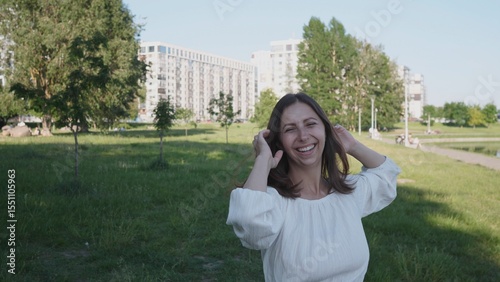 A woman stands in a park, smiling happily while playing with her hair. Modern apartment buildings are visible in the background, highlighting an urban setting.