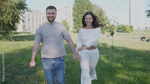 Two people happily stroll together in a park, holding hands and smiling as they enjoy a sunny day surrounded by nature and contemporary architecture.