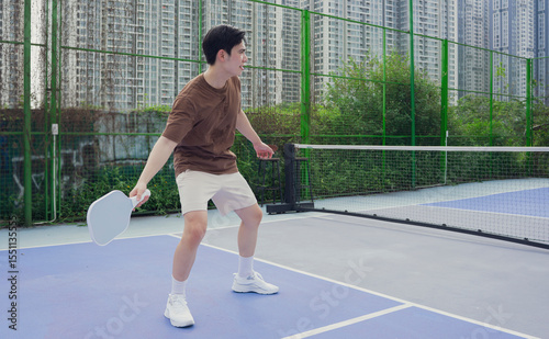 Man plays pickleball on an outdoor court surrounded by urban high-rise buildings, enjoying a sunny day with friends