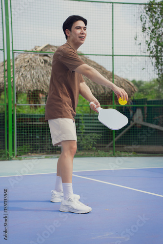 Young Asian man enjoys pickleball on a sunny day at a vibrant outdoor court surrounded by greenery