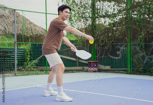 Young man plays pickleball on outdoor court surrounded by greenery during sunny day in Asian community setting