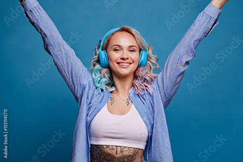 Young woman casual clothes listen to music in headphones dance raise up hands isolated on plain blue background studio. Lifestyle portrait