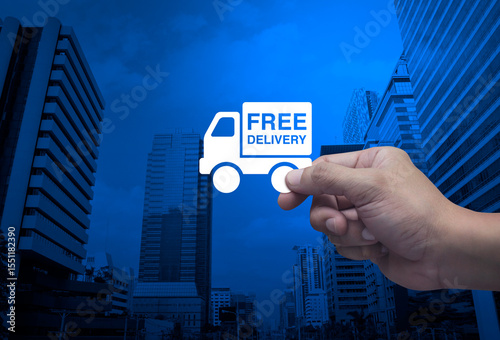 Hand holding free delivery truck flat icon in fingers over modern office city tower and skyscraper, Business transportation service concept