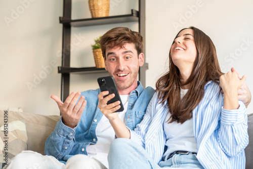 Happy young couple sitting on the sofa at home celebrating Happy young couple sitting on the sofa at home celebrating after checking great news on a smartphone. Passed exam, driving test, school resul