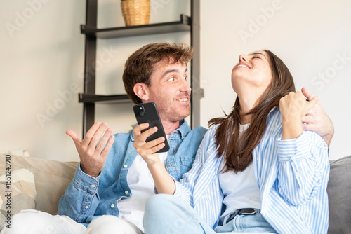 Happy young couple sitting on the sofa at home celebrating Happy young couple sitting on the sofa at home celebrating after checking great news on a smartphone. Passed exam, driving test, school resul