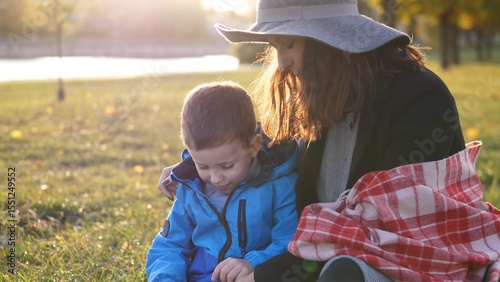 A mother and her young son sit together in a park, enjoying the sunshine and exploring the grass as autumn leaves decorate the surroundings, creating a cozy atmosphere.