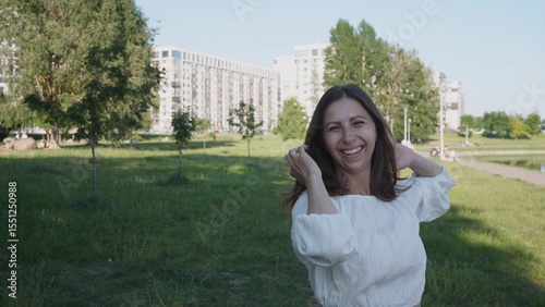 A cheerful woman stands in a lush park, playfully touching her hair while showcasing her smile against a backdrop of urban architecture and greenery.