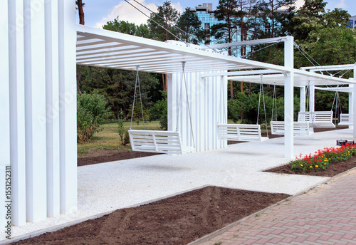 A modern outdoor space featuring white swings under a minimalist structure. The area is surrounded by greenery and flower beds