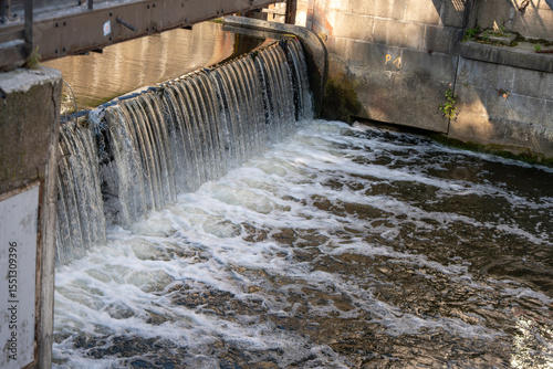 A small waterfall flows over a weir into a river. The structure demonstrates the combination of industrial construction and flowing water.
