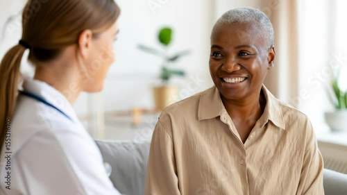  A cheerful senior African American woman smiles while talking with her female doctor during a home visit. Representing positive patient care and supportive senior healthcare.