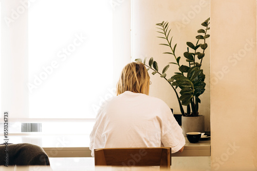 Woman working at desk by window with potted plant and coffee cup, copy space.