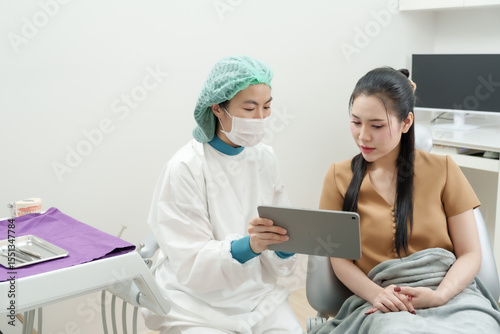 asian male dentist wearing surgical cap mask gown using digital tablet to explain root canal treatment to adult asian female patient sitting on dental chair in modern dental clinic