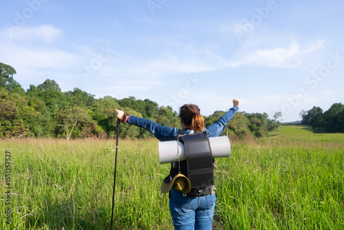 Rear view of an Asian woman with a solar-powered backpack raising her arms, symbolizing sustainable hiking, off-grid adventure, and eco-friendly travel in nature.