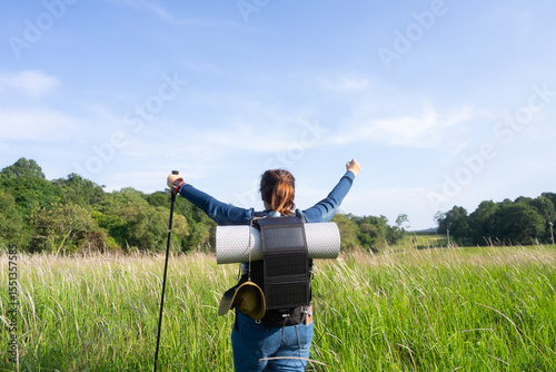 Rear view of an Asian woman with a solar-powered backpack raising her arms, symbolizing sustainable hiking, off-grid adventure, and eco-friendly travel in nature.