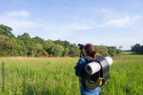 An Asian woman uses binoculars to explore the forest, carrying a backpack equipped with a solar panel charger, symbolizing sustainable hiking, off-grid adventure, and eco-friendly travel.