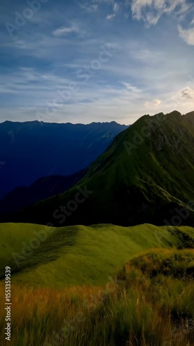 Picturesque mountain valley with grassy hillside leading to towering peaks and radiant sunlight breaking through dramatic clouds