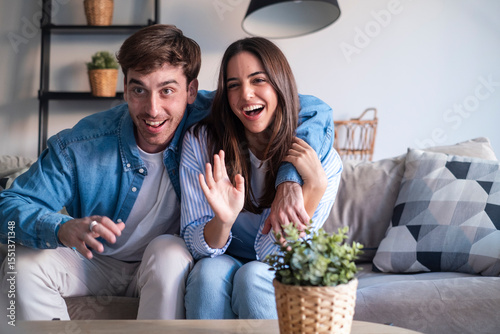 Happy young couple in a video call from home, smiling and sharing joyful news through the laptop or phone camera. Announcing engagement, vacation plans, or exciting updates with friends or family