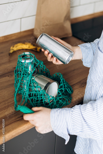 Woman putting empty metal cans into mesh bag in kitchen, recycling concept.