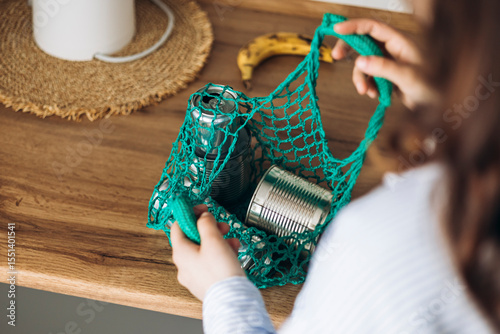 Woman putting empty metal cans into mesh bag in kitchen, recycling concept.