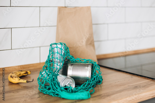 Mesh bag with cans, paper bag, and rotten banana on kitchen table, waste.