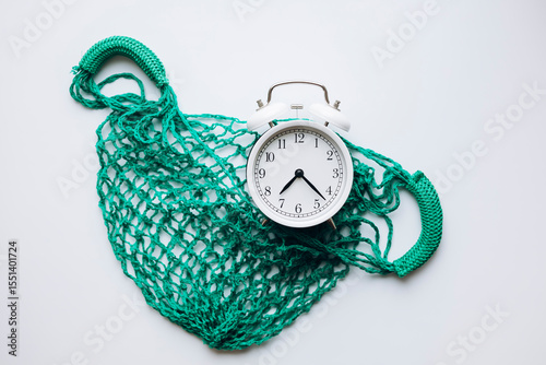 Flat lay with green mesh bag and white alarm clock on clean white background.