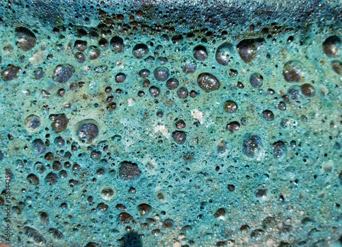 A macro shot reveals an abstract, porous surface with a vibrant turquoise glaze, featuring numerous raised bubbles and dark, pitted depressions, creating a highly textured pattern