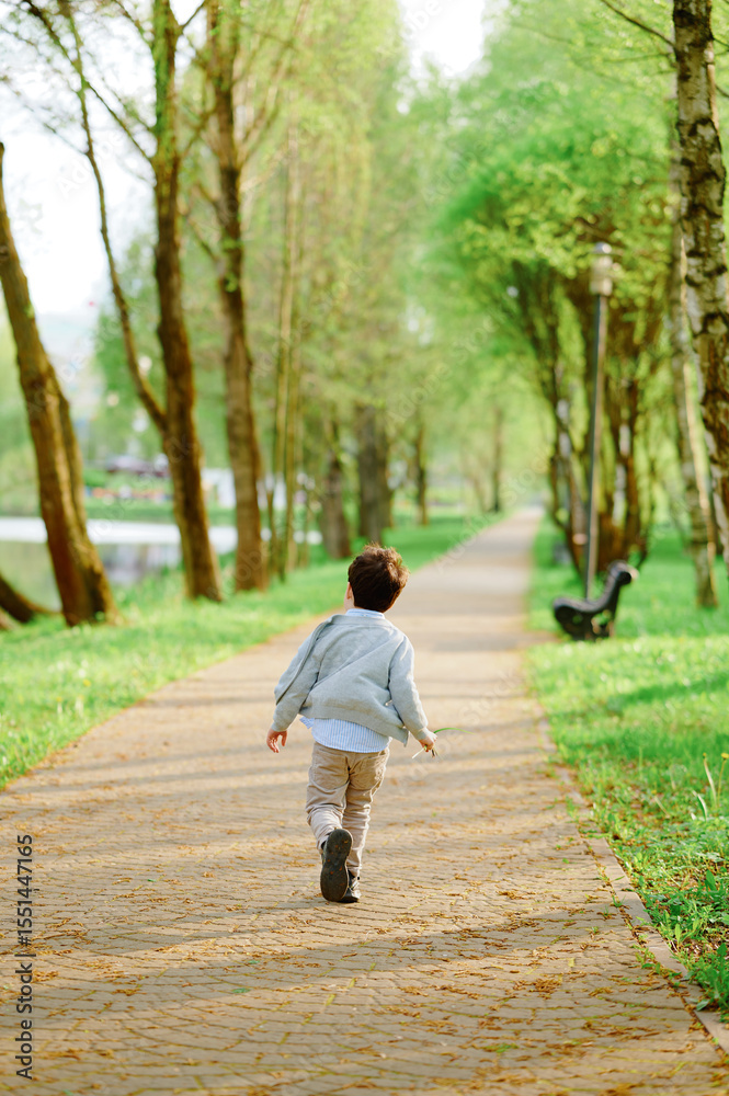 Fototapeta premium child walking on park pathway surrounded by lush green trees and sunlight. casual outfit suggests relaxed outdoor activity. perfect for concepts of childhood, exploration, nature, innocence