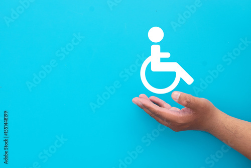 A hand supporting a wheelchair user icon.Concept for supporting people with disabilities, disability rights, inclusion,accessibility,and providing care and assistance.Blue background with copy space.