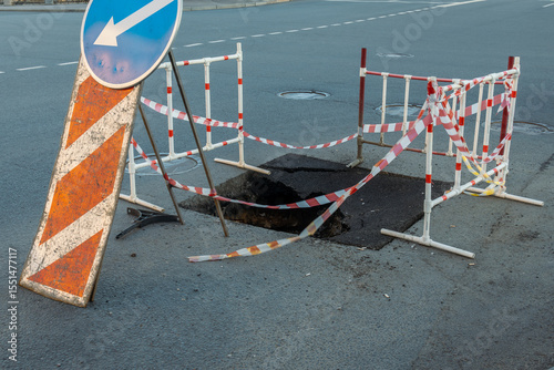 Blue arrow pointing to the left is on a sign. The sign is next to a road with a hole in it. There are orange and white caution tape and barricades around the hole