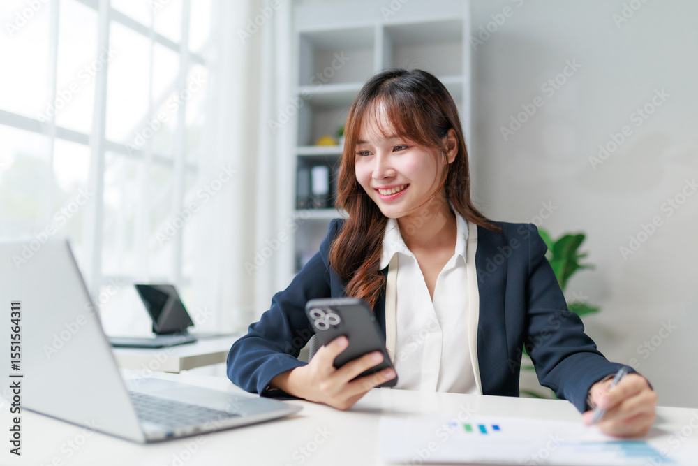 Young Asian businesswoman holding smartphone and working with financial documents, marketing graphs, and laptop at office desk Young Asian businesswoman holding smartphone and working with financial documents, marketing graphs, and laptop at office desk
