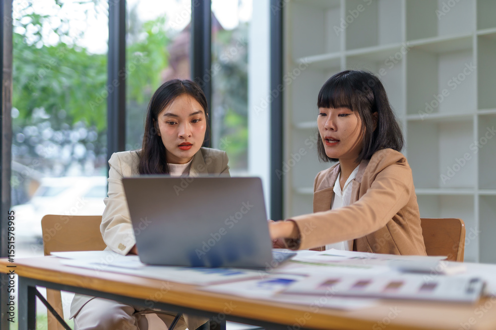 Fototapeta premium Two Asian businesswomen collaborating and discussing strategies while working on a laptop in a modern office environment