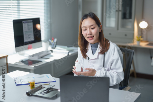 Asian female doctor wearing lab coat holding medicine bottle having video conference with patient on laptop in hospital office
