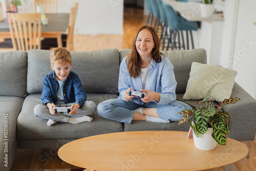 Wallpaper Mural A cheerful mother and her young son sitting on a cozy couch, fully immersed in playing video games together. Torontodigital.ca