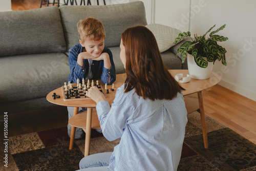 Wallpaper Mural Happy mother playing chess with her 7-year-old son at home. Concept of childhood development, quality time, strategy, and joyful parenting in a cozy home environment. Torontodigital.ca