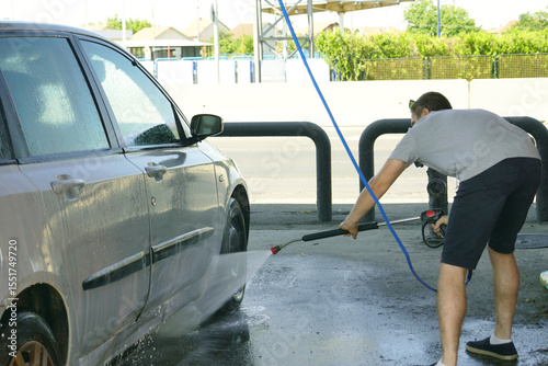 Rear view of adult man focused on car washing task using hose with water spray at urban car wash. Masculinity, care for possessions, focus on task, and modern man image in everyday context.