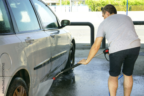 A young man bends over and washes the underside of a silver-gray passenger car with a high-pressure water gun at a self-service car wash in an open-type bay. Self-cleaning of personal transport