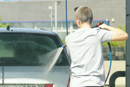 Young man using high-pressure water spray to wash passenger car at self-service car wash station. Concept of independence, daily chores, car care routine, water cleaning and personal responsibility