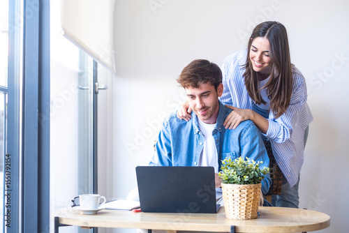 Happy young couple sitting at home together, smiling while looking at laptop and documents. Positive moment of planning, talking to friends or family, or enjoying success in work, finances or future p
