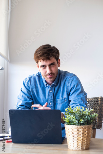 Young man in blue shirt sitting at a table with laptop, thoughtful but calm during a work break at home or in a hotel. Modern remote professional lifestyle, digital tools, and flexible working habits