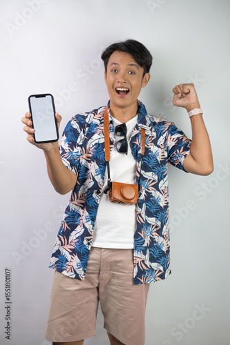 Happy young asian man wearing blue summer shirt is holding a smartphone with blank screen for mock up mockup, isolated over white background.